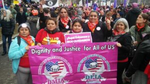 Picture of women marching at the head of a crowd carrying a sign that says, "Dignity and Justice for all of us"