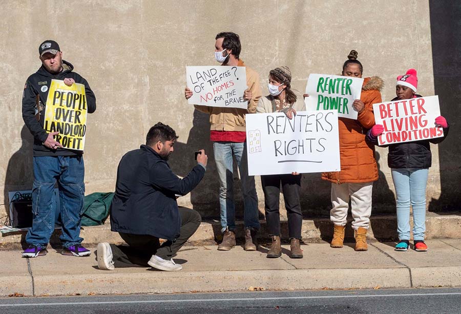 Photo of five members standing in front of a wall holding signs about tenant rights. A man takes video of them.