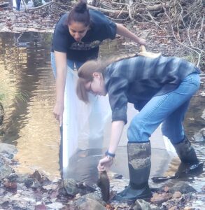 Two women work with a plastic strainer at the edge of a river