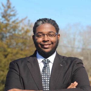 Picture of Justin Nick, a Black man in a gray suit standing outside in front of trees.