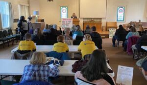 Picture of a worship space with a woman behind the podium and a large crowd behind tables