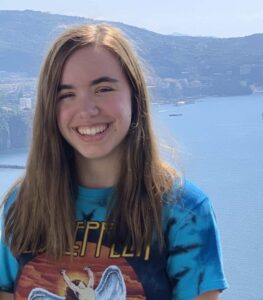 Profile picture of a young white woman with brown hair and eyes and a big smile standing in front of a landscape with a lake or bay and mountains in the background.
