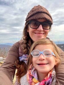 Picture of a woman and child outside with gray sky above. Both wear glasses and smile.