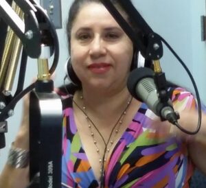 Picture of a Latina woman in a studio with a big microphone. She is wearing a colorful dress and lipstick.