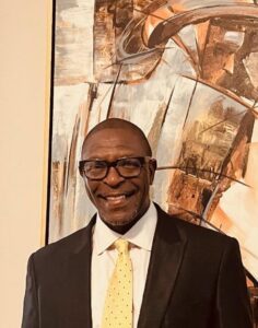 Image of a Black man wearing glasses and a business jacket and tie standing in front of a modern cubist style of painting in shades of brown and white