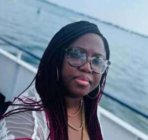 Photo of a Black woman with glasses and long braids standing on a boat at the edge of water.