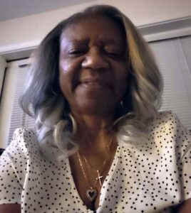 Selfie of a Black woman with gray hair smiling, wearing a nurse's white and black polka dot shirt, and gold necklaces with heart and cross pendants.