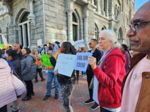 Picture of a diverse crowd of people standing outside a church in downtown Richmond holding signs. One white woman with white hair holds a sign that reads, "End Jim Crow."