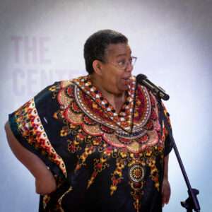 Picture of a Black woman with short gray hair and glasses and a very colorful shirt speaking into a microphone