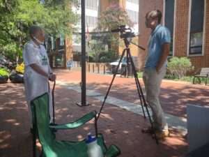 Picture of a white woman with gray hair being interviewed by a white man with a camera on the downtown mall in Charlottesville
