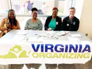 Picture of 4 people sitting behind a table, one holds a dog, with a Virginia Organizing tablecloth with logo in front of them and windows behind